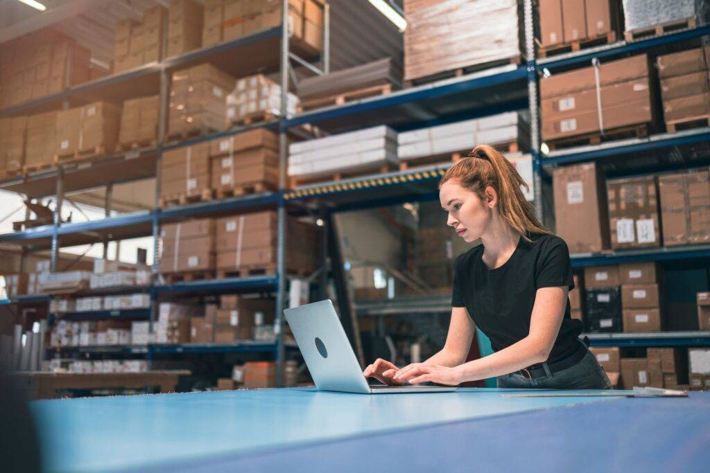 Eine junge Frau arbeitet in einem Lager an ihrem Laptop, um die Lagerverwaltung und E-Commerce-Prozesse zu optimieren, unterstützt durch eine Amazon Agentur.