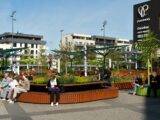 Menschen sitzen auf modernen Parkbaenken aus Holz rund um bepflanzte Inseln auf einem Platz in einem Stadtquartier