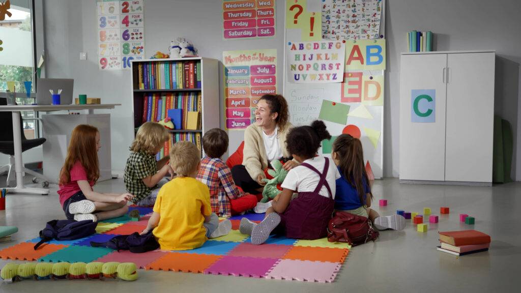 Kindergartengruppe sitzt mit einer Erzieherin auf bunten Matten im Klassenraum – zeigt den Alltag und die Chancen fuer Quereinsteiger Kindergarten in der fruehkindlichen Bildung.